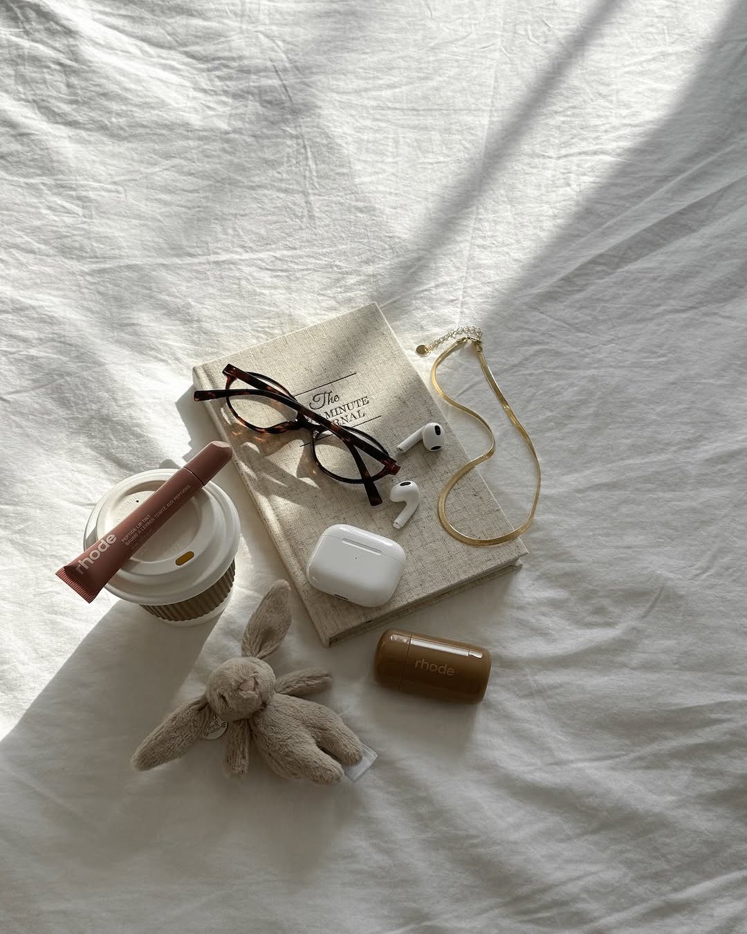 A flat lay of items on a white bed, perfect for an after-school routine: a book, eyeglasses, gold necklace, AirPods, to-go coffee cup, lip balm, brown container, and a small plush bunny toy in soft sunlight.