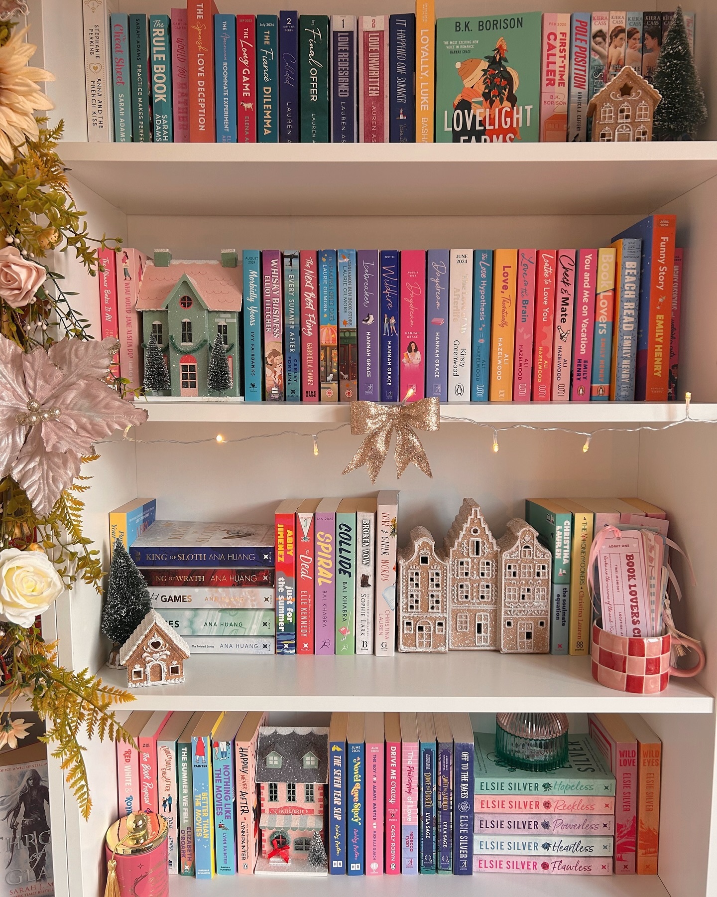 A white bookshelf decorated with mini houses, fairy lights, and flowers. The shelves hold colorful books, mostly romance novels, arranged both vertically and horizontally. Festive decorations and mugs add cozy charm.