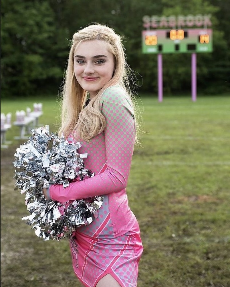 A smiling cheerleader with long blonde hair, channeling Disney Stars energy, holds silver pom-poms and wears a pink and gray uniform, standing on grass with a scoreboard and benches in the background.