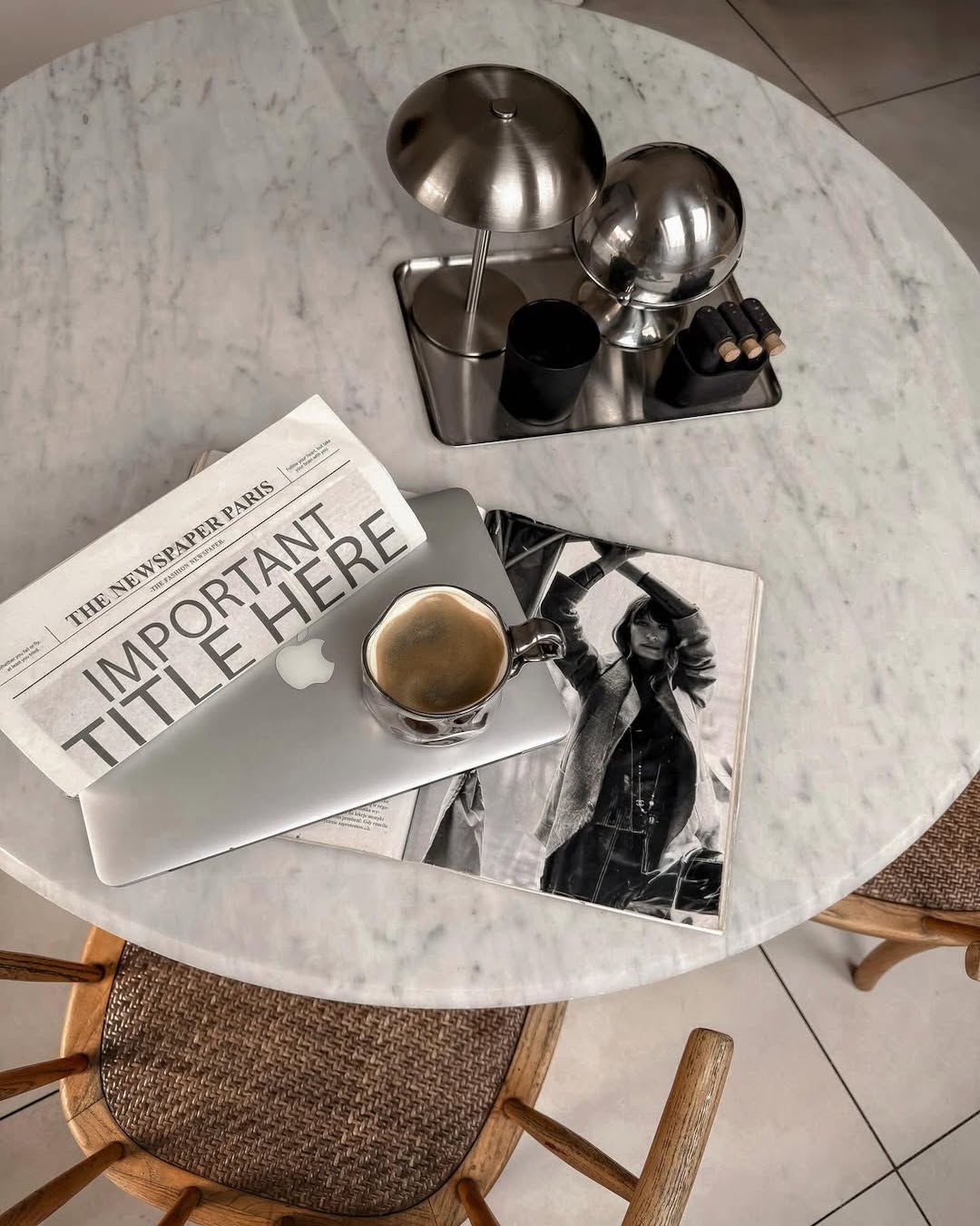 A marble table set for the best after-school routine, with a laptop, coffee cup, newspaper, magazine, silver lamp, black cup, and a set of round objects. Two wooden chairs are partially visible.