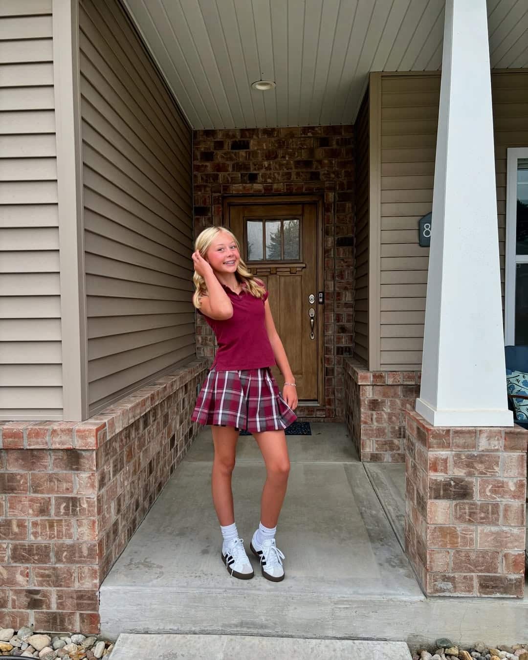 A girl in a burgundy top and plaid skirt poses on the front porch, smiling with one hand in her hair. She sports white sneakers, embodying the best after-school routine as she stands on a concrete walkway framed by brick pillars.