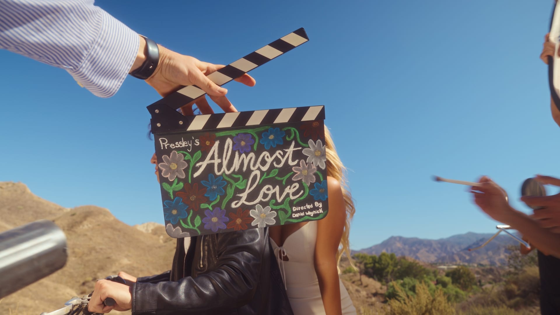A hand holds a colorful clapperboard labeled Pressley's Almost Love, in front of two people outdoors on a sunny day with mountains and a bright blue sky in the background.