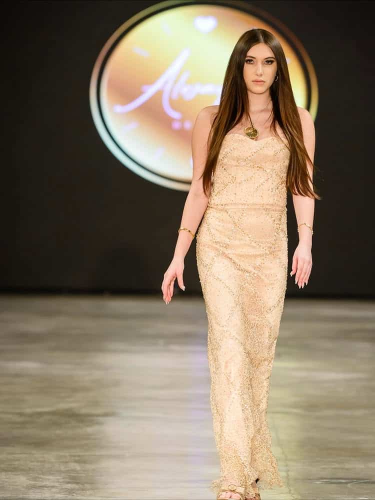 A 15-year-old female model with long brown hair walks a runway in a gold, sparkling sleeveless gown. The background features an exclusive circular, blurred logo and a smooth, polished floor, showcasing the work of a talented fashion designer.