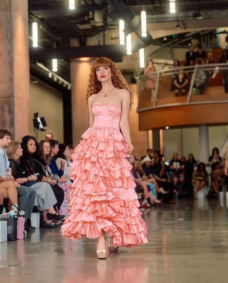 A model with long red hair walks the runway in a strapless, pink, ruffled dress by a 15-year-old fashion designer. She strides under bright lights before seated spectators and more fans watching from the balcony above.