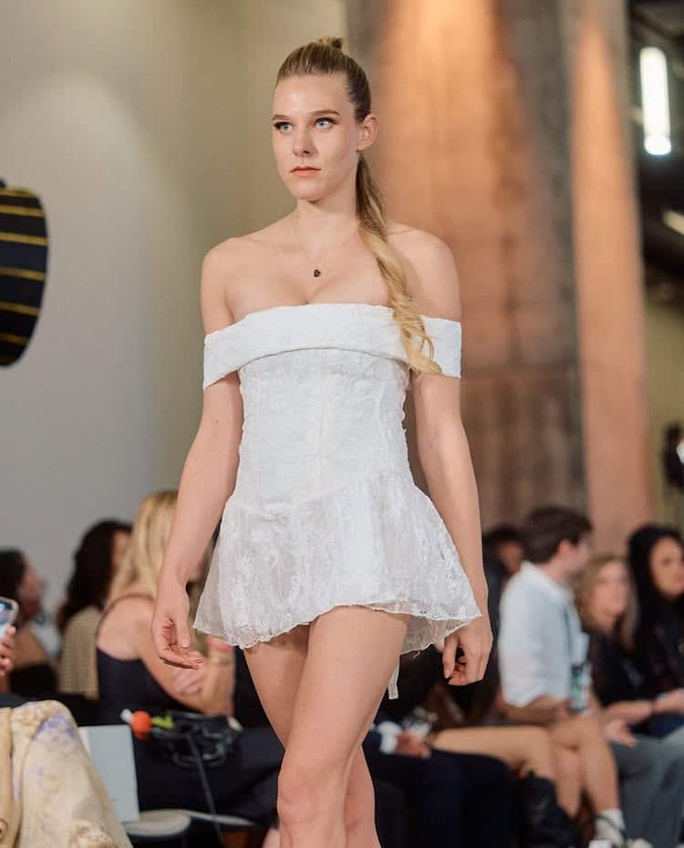 A model with long blonde hair in a ponytail walks a runway wearing an exclusive off-the-shoulder, short white lace dress. An audience sits in the background, watching the fashion show.