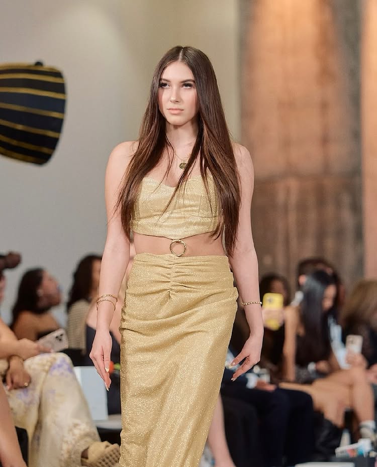 A female model with long brown hair walks on a runway in a shimmering gold two-piece outfit by an exclusive 15-Year-Old Fashion Designer, surrounded by an audience seated and taking photos at a fashion event.
