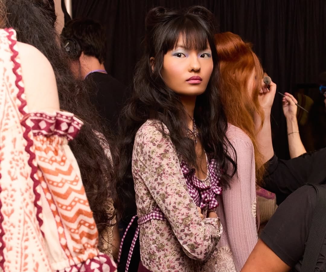 A young woman with long dark hair styled in buns and waves stands among others. Radiating full-glam inspo, she wears a floral patterned dress with a purple gingham bow and bold blue eyeshadow, looking thoughtfully to the side.