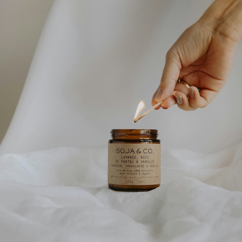 A hand lights a brown glass candle labeled “SOJA & CO. Lavande, Bois de Santal & Vanille” with a match, against a soft, white fabric background—perfect for unwinding after a long day of debunking wellness myths on the internet.