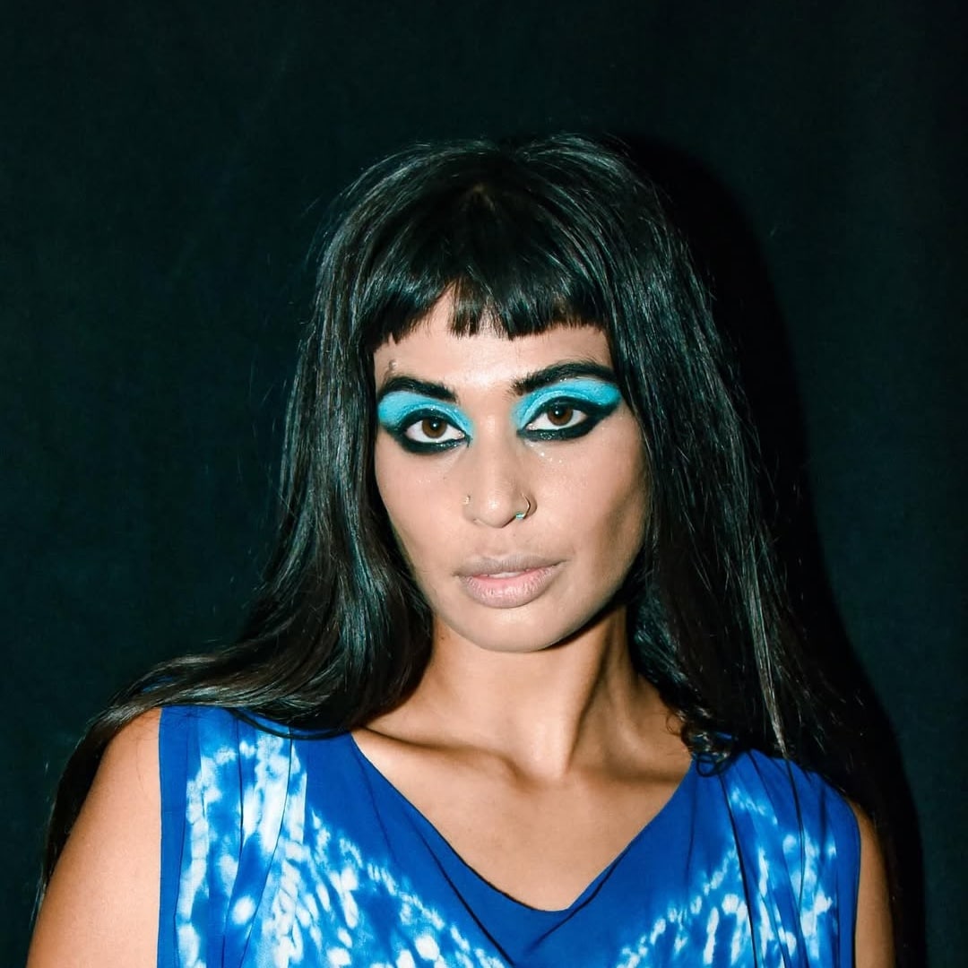 A person with long dark hair and bold blue eyeshadow and eyeliner stands against a black background, channeling full-glam inspo in a blue patterned top, looking directly at the camera with a neutral expression.