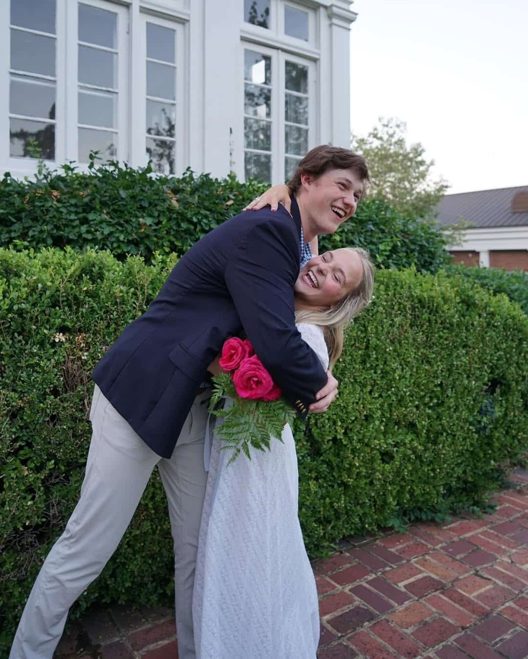 A young man and woman hug and smile joyfully outside in front of a hedge and windowed building. The woman holds three pink roses—a sweet result of his courage to ask out your crush—while she wears a white dress and he a dark blazer.