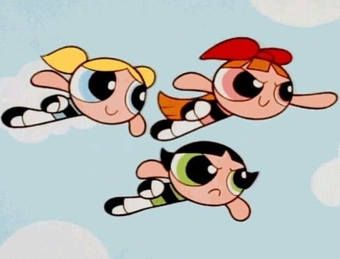 Three animated superhero girls with large eyes and colorful outfits—one with blonde hair, one with orange hair, and one with black hair—are flying through a blue sky with clouds.
