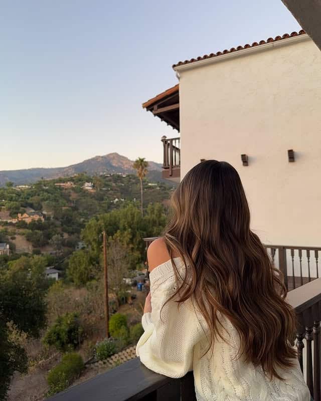 A woman with long wavy brown hair, wearing an off-shoulder white sweater, stands on a balcony at sunset, gazing at scenic hills and greenery—much like a scene with the PJO season 3 characters cast enjoying some peace.