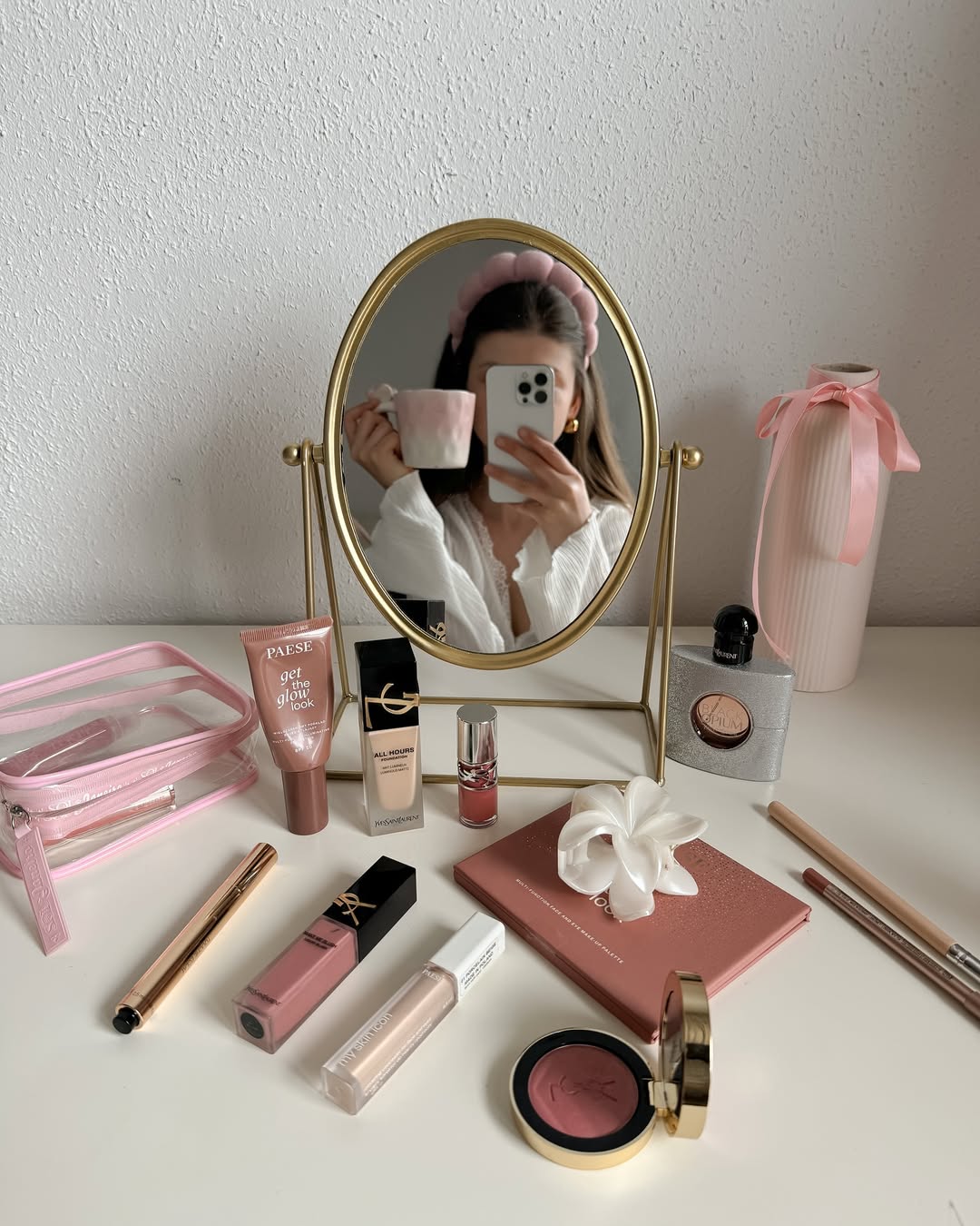 A woman in a white top, seen in a round gold mirror, holds a pink mug and phone. Surrounded by style tips essentials—a pink pouch, scrunchie, and makeup—she shows how to feel put-together even when in a rush. The setting is bright and tidy.
