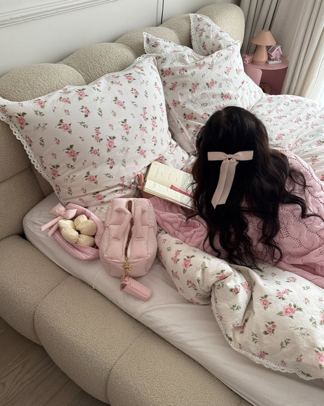 A person with long dark hair and a pink bow reads a book on a bed with white and pink floral bedding. A pink bag, braided snack, and cozy decor create a soft vibe—perfect for learning how to feel put-together even in a rush.