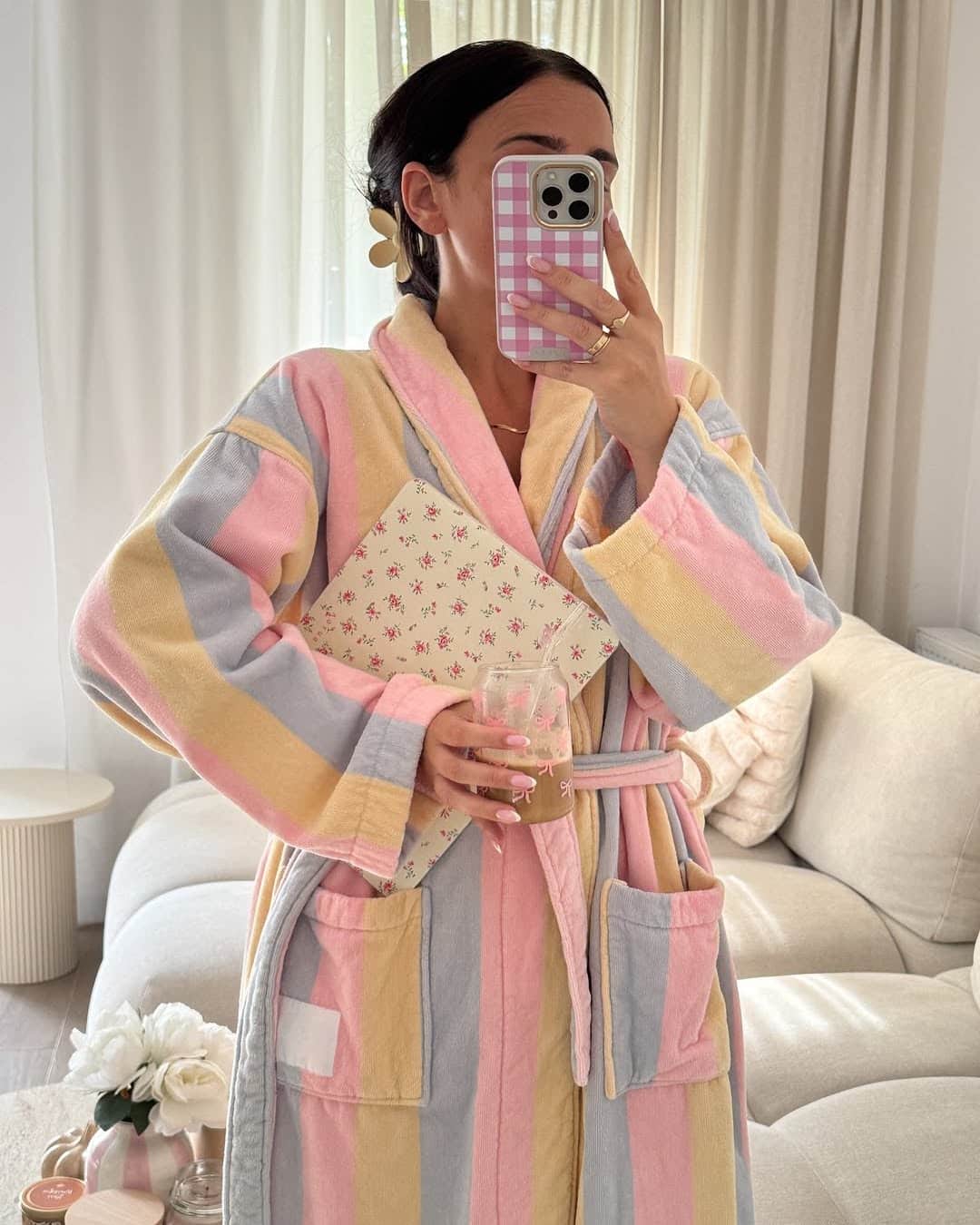 A person in a pastel striped robe holds a floral notebook and a glass of iced coffee, taking a mirror selfie with a phone in a pink checkered case. Even in a rush, they look effortlessly put-together in their cozy living room with light furnishings.