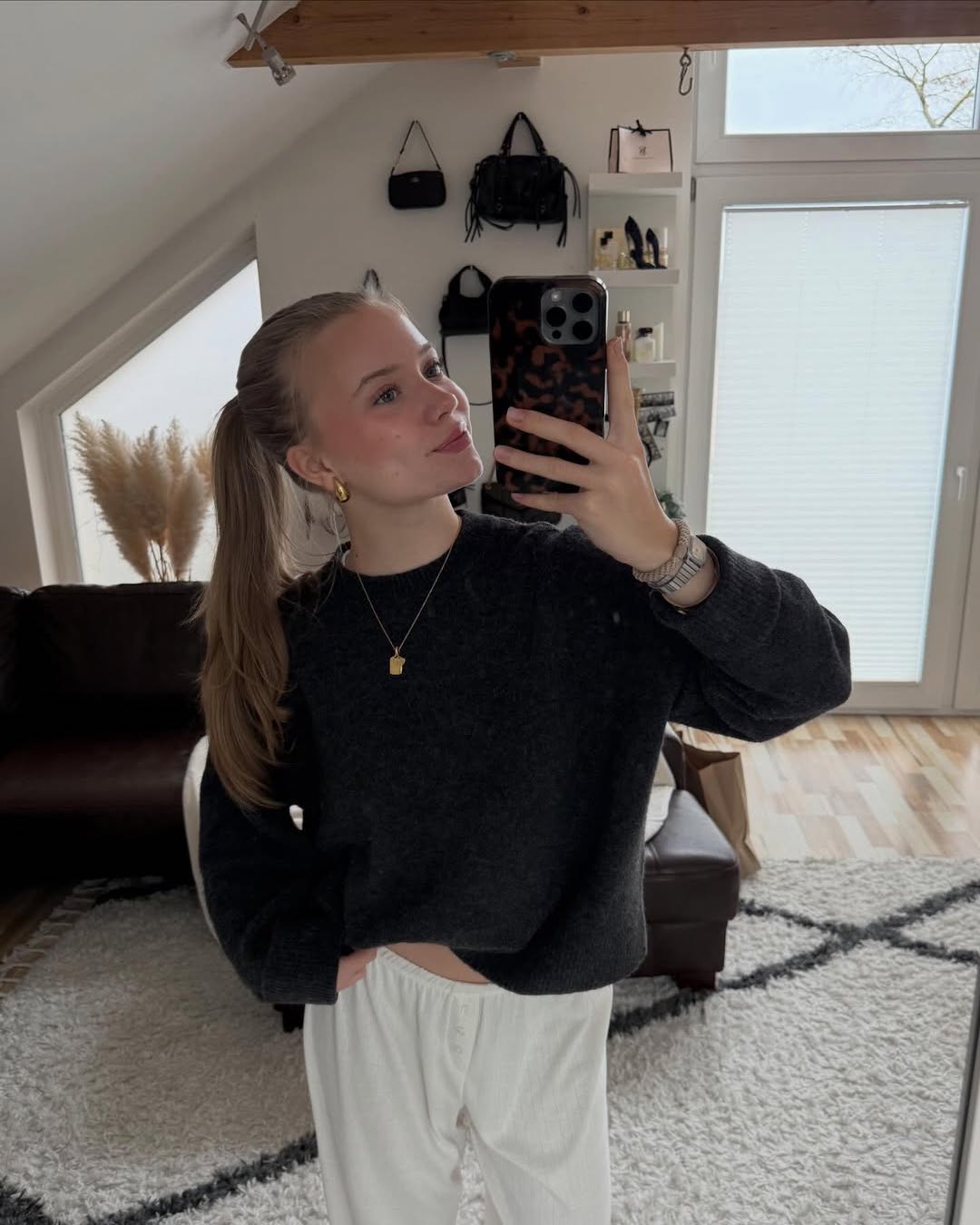 A young woman with a ponytail takes a mirror selfie in a modern, light-filled room. She wears a dark sweater, white pants, and gold jewelry. The room has a couch, shelves with bags, and a large window with blinds.