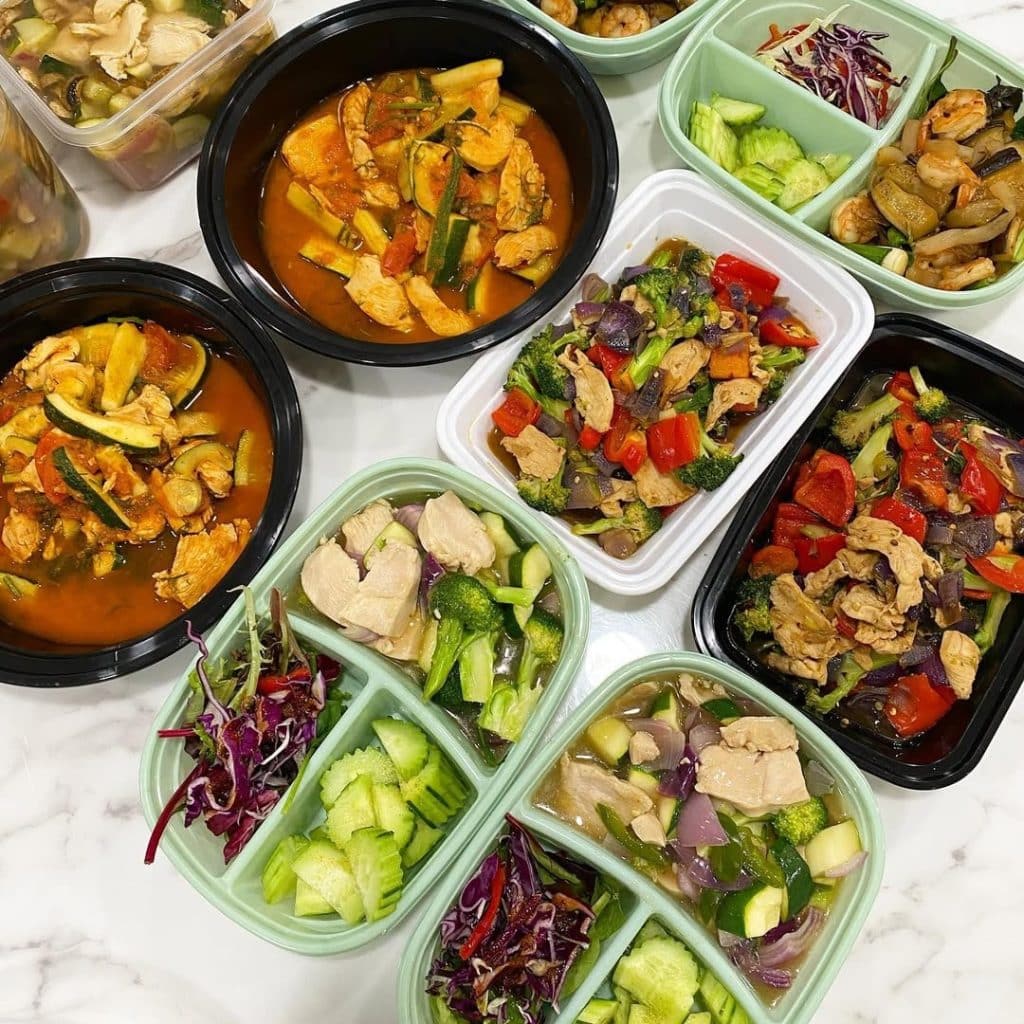 Several meal prep containers filled with colorful dishes, including stir-fried chicken with vegetables, cucumber slices, salad greens, and red cabbage—perfect for anyone looking for a beginners guide to meal prepping—are arranged neatly on a white marble surface.