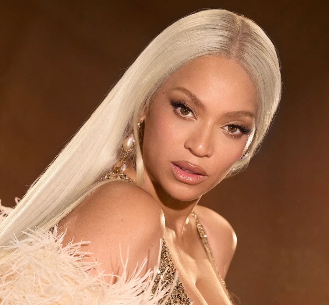 A woman with long, straight platinum blonde hair poses in a glamorous, gold outfit with feathered details—full-glam inspo that channels The Life of a Showgirl—against a warm brown background, looking confidently at the camera.