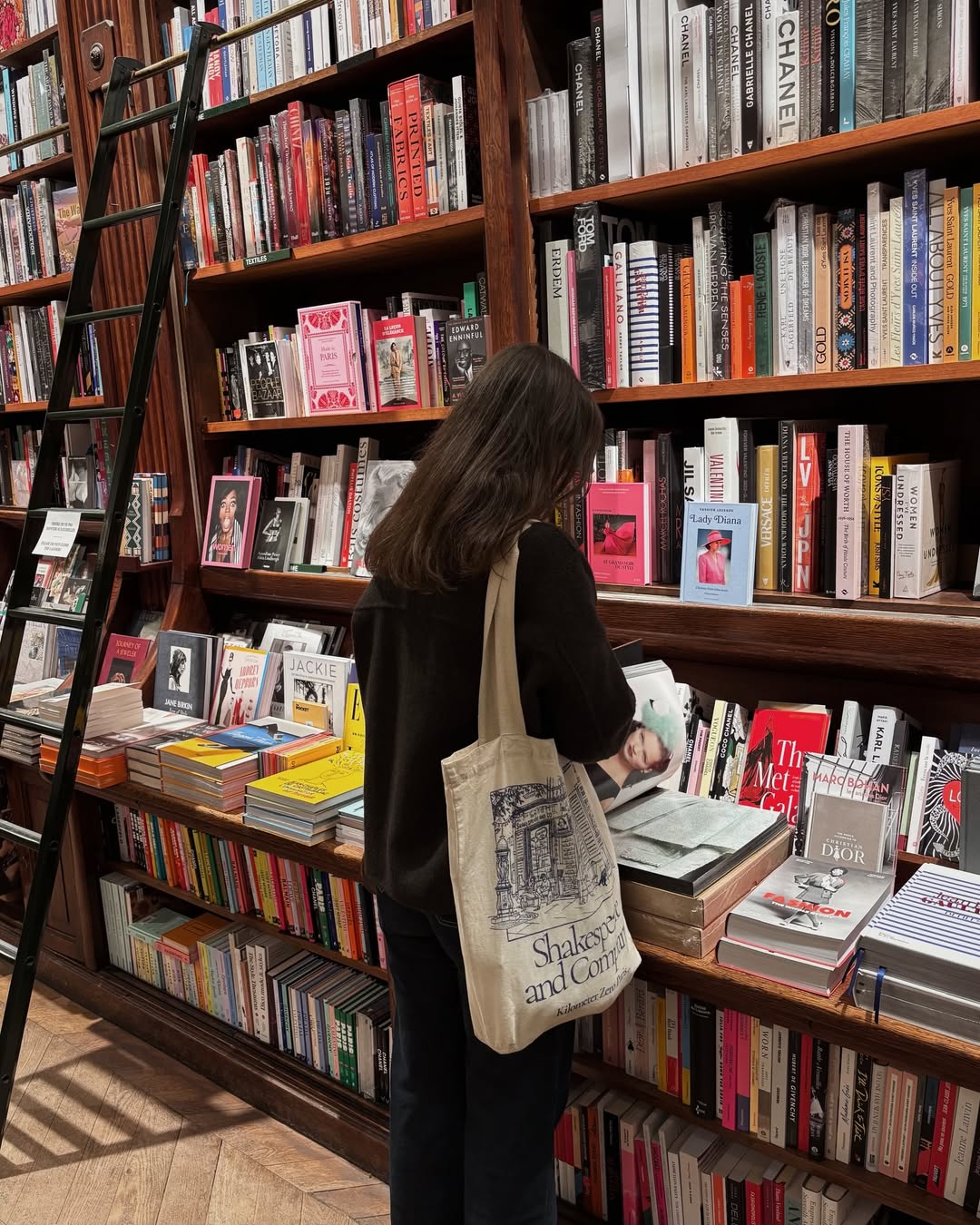 A person with long brown hair stands in front of bookshelves, browsing a book. They wear dark clothes and carry a beige Shakespeare and Company tote bag. The shelves are filled with books, and a ladder leans against them.