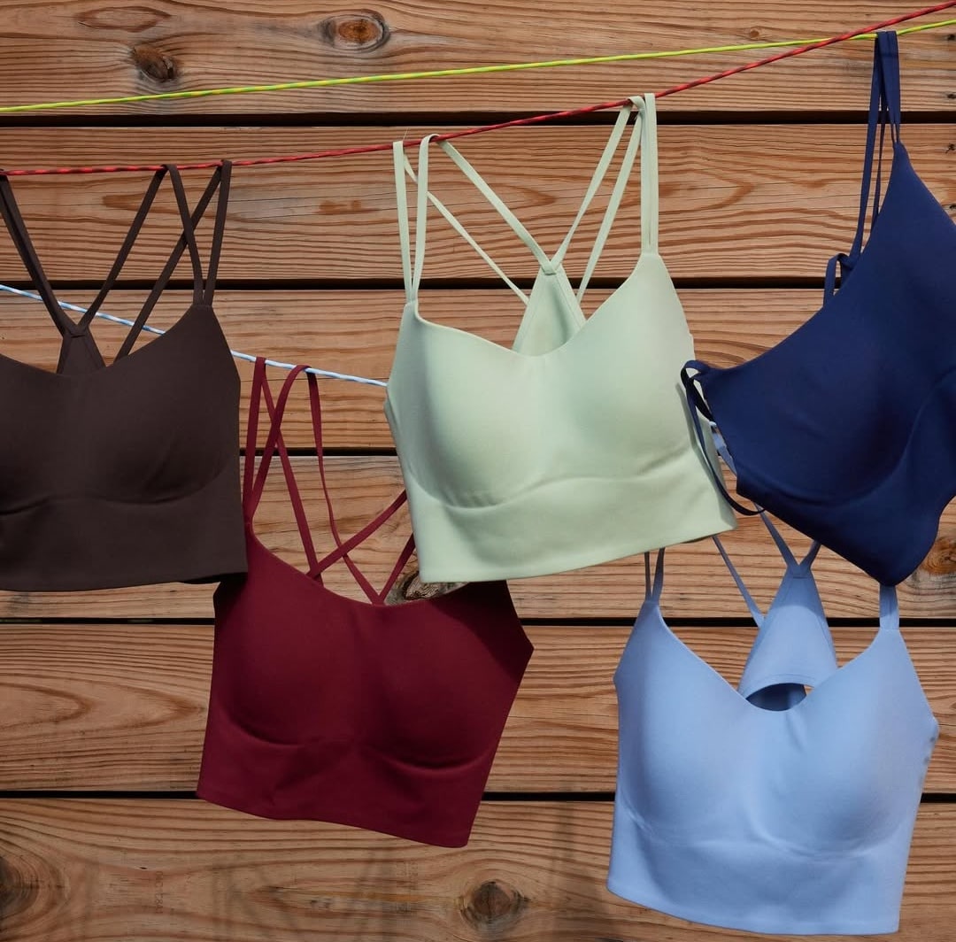 Five strappy bralettes in brown, maroon, cream, navy blue, and light blue hang from colorful clotheslines in front of a wooden plank wall—a playful snapshot for your perfect bra fit or next bra guide.