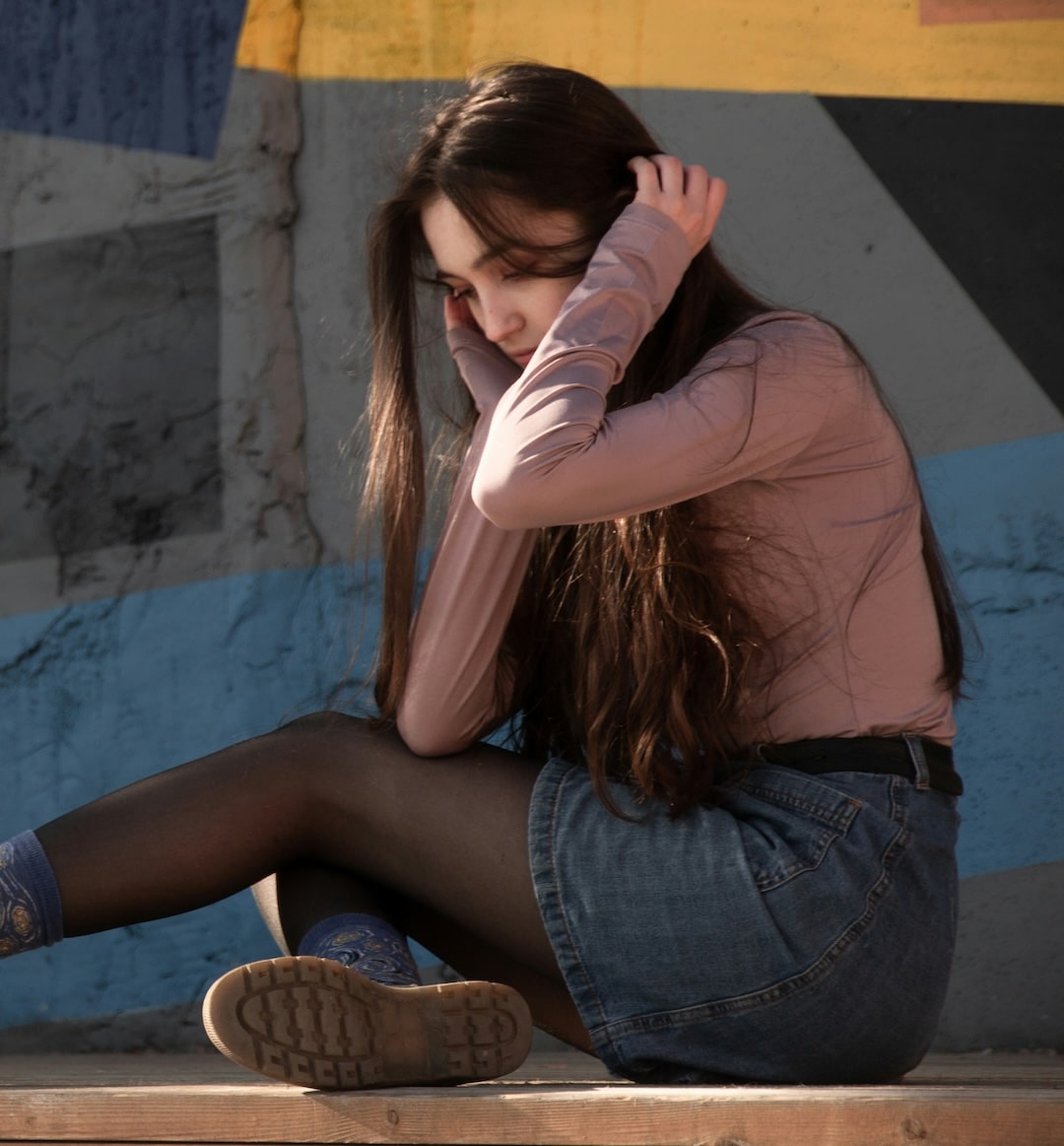 A young woman with long brown hair sits on a wooden surface, resting her head on her hand, as if contemplating how to break up. She wears a mauve long-sleeve top, denim skirt, black tights, and blue socks. A colorful wall is in the background.