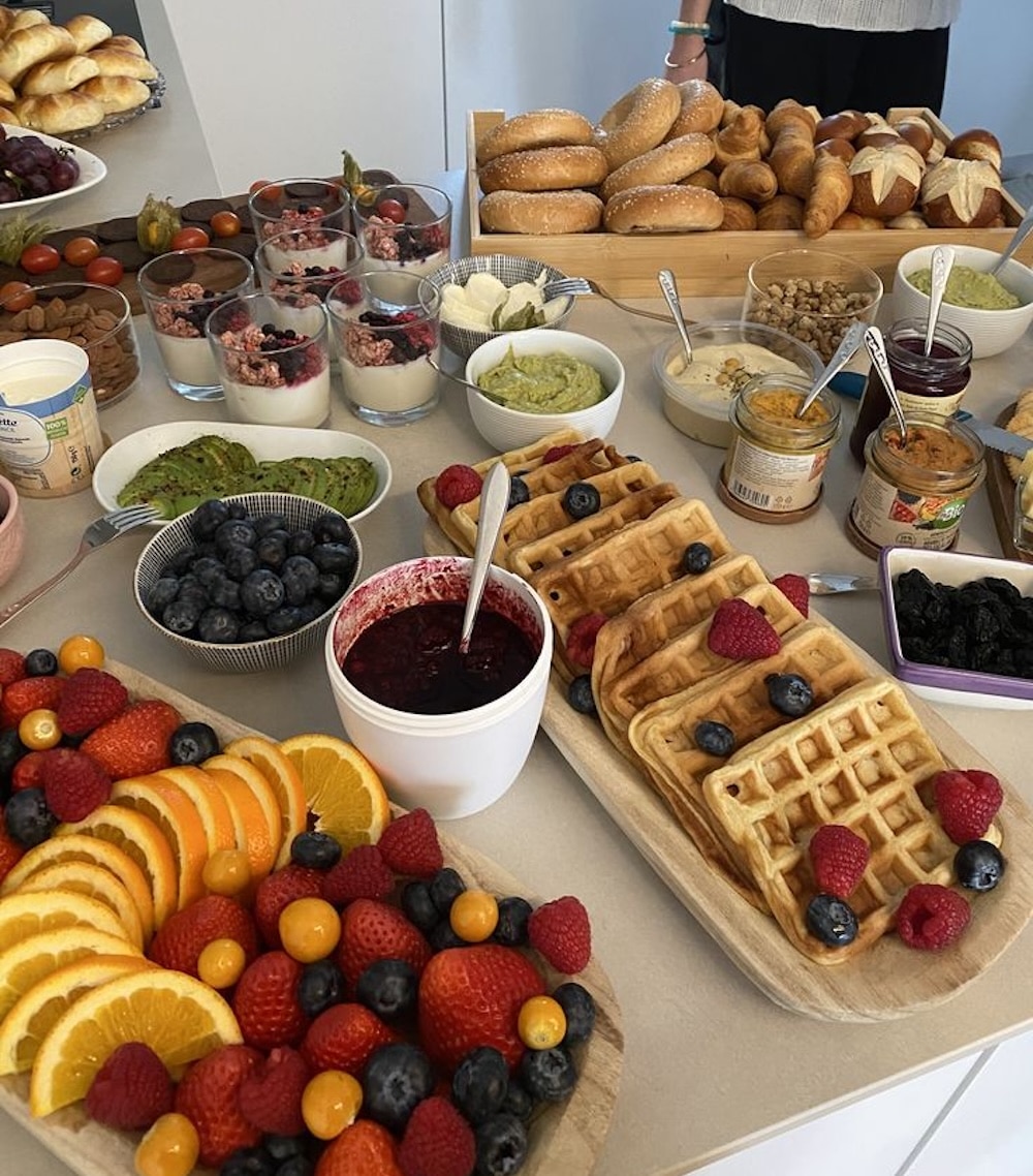 A perfect Spring brunch spread awaits as you host your squad: waffles topped with berries, bowls of fruit, sliced oranges, bread rolls, yogurt parfaits, dips, spreads, and assorted toppings all arranged on a counter.