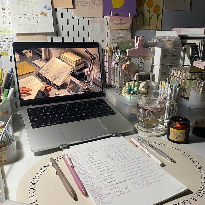 A neatly organized study desk with a laptop, handwritten notes, pens, highlighters, school supplies, a clear mug with a drink, a lit candle, stationery items, and decorative items against a pegboard and wall with various papers.