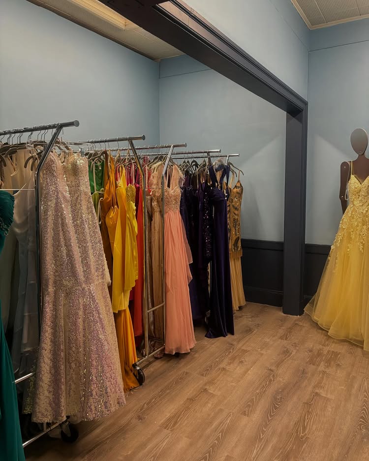A room with several racks of colorful formal dresses, including pastel, yellow, orange, and dark shades. A mannequin displays a yellow gown on the right. The floor is wooden and the walls are light blue—sustainable fashion at its finest.
