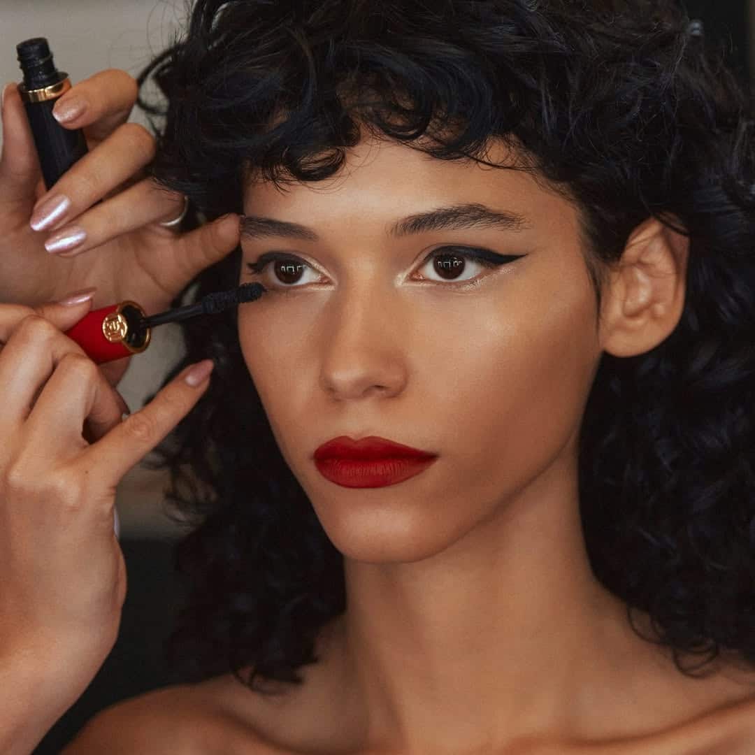 A woman with curly dark hair and bold red lipstick gets black mascara applied to her lashes, channeling full-glam inspo. She wears winged eyeliner and gazes slightly to the side, evoking the allure of The Life of a Showgirl.