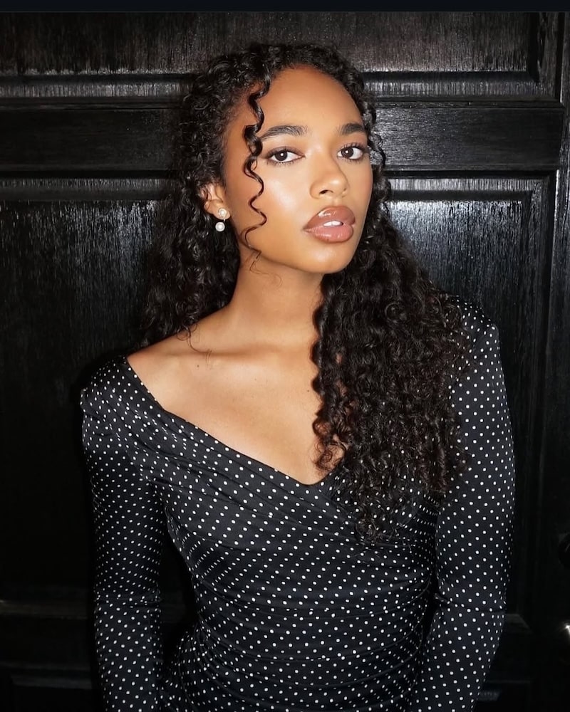 Channeling Elle’s signature confidence from the Legally Blonde series, a woman with long curly hair in a black dress with white polka dots stands in front of a dark wooden background, looking boldly at the camera.