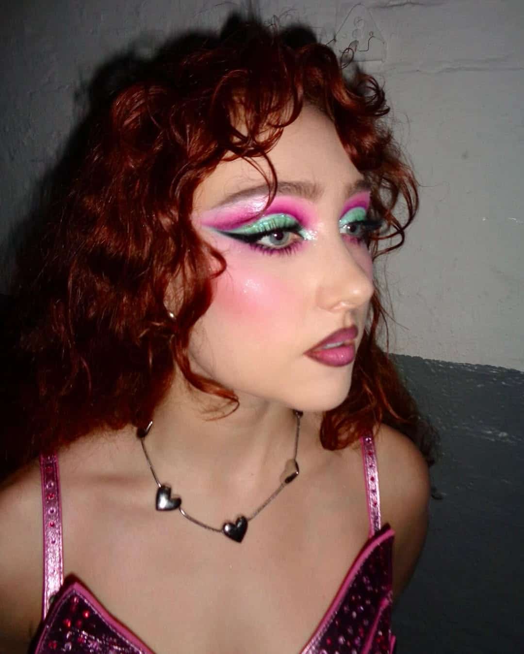 A person with curly red hair wears full-glam, colorful makeup—dramatic pink and green eyeshadow, glitter, and blush. With dark lipstick, a pink outfit, and a heart necklace, they channel showgirl vibes while standing against a plain wall.