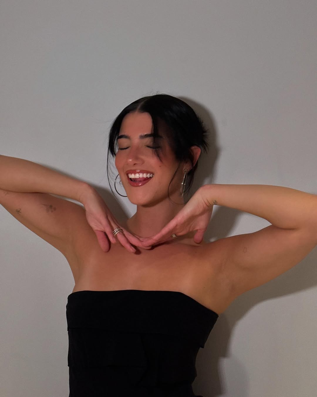 A woman in a black strapless top smiles with her eyes closed, framing her face with both hands under her chin. She has dark hair tied back and wears large hoop earrings—a joyful pose that perfectly captures simple beauty tips in action.