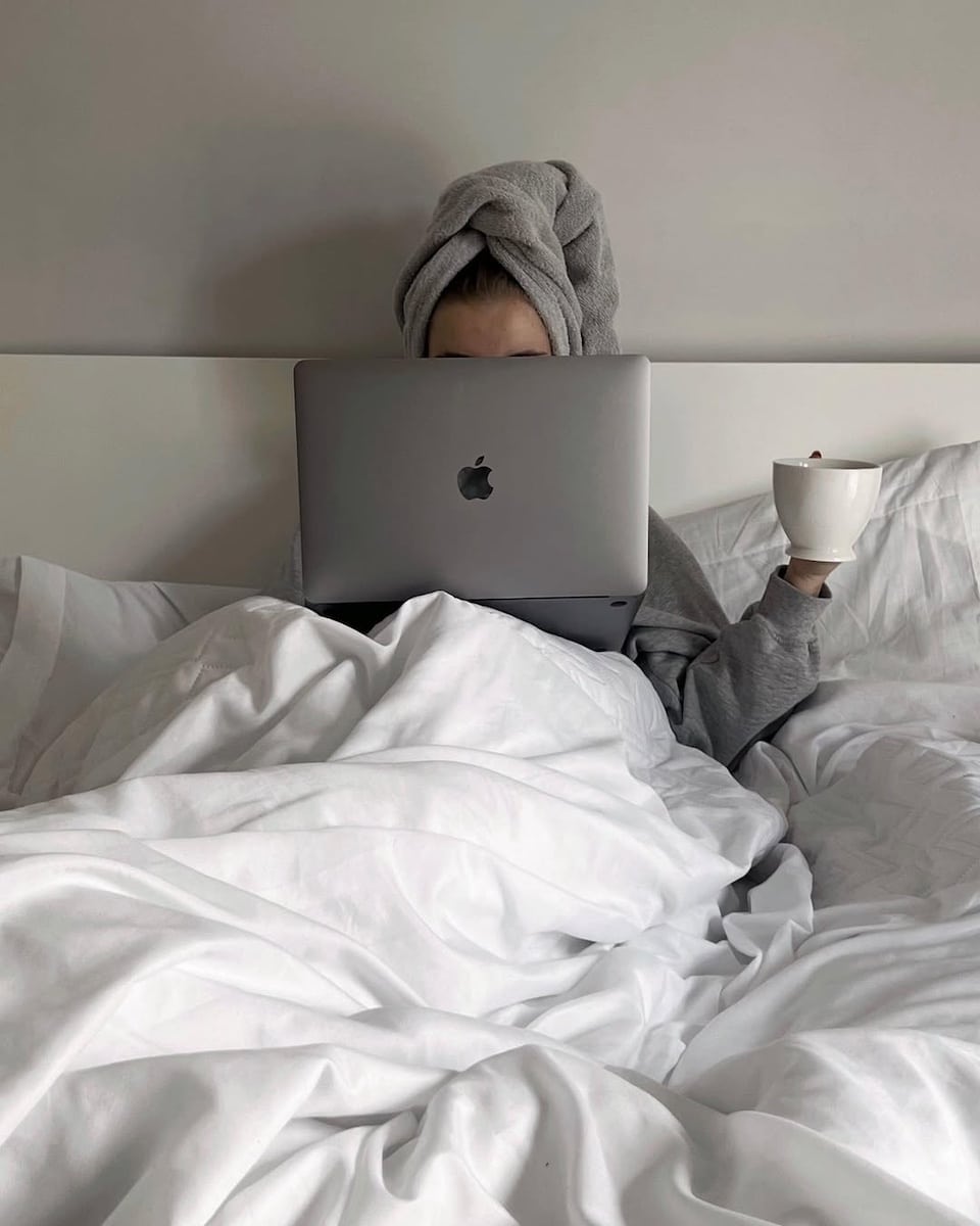 A person sits in bed under white blankets, wearing a gray towel wrapped around their head, holding a white mug and using a laptop with the Apple logo visible, ready to open ChatGPT for a cozy online session.