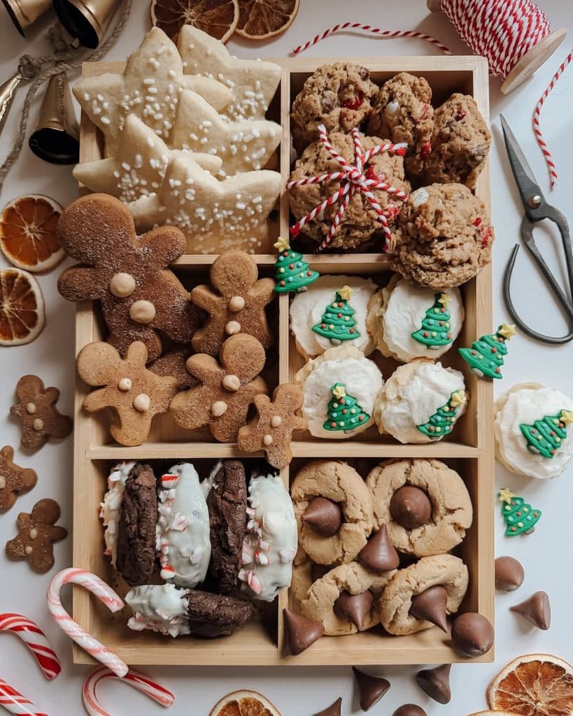 A wooden box divided into six sections, each filled with assorted Christmas cookies, including gingerbread people, frosted sugar cookies, tree-shaped cookies, and chocolate cookie favorites. Candy canes and dried orange slices add festive charm—perfect for a Christmas quiz night.