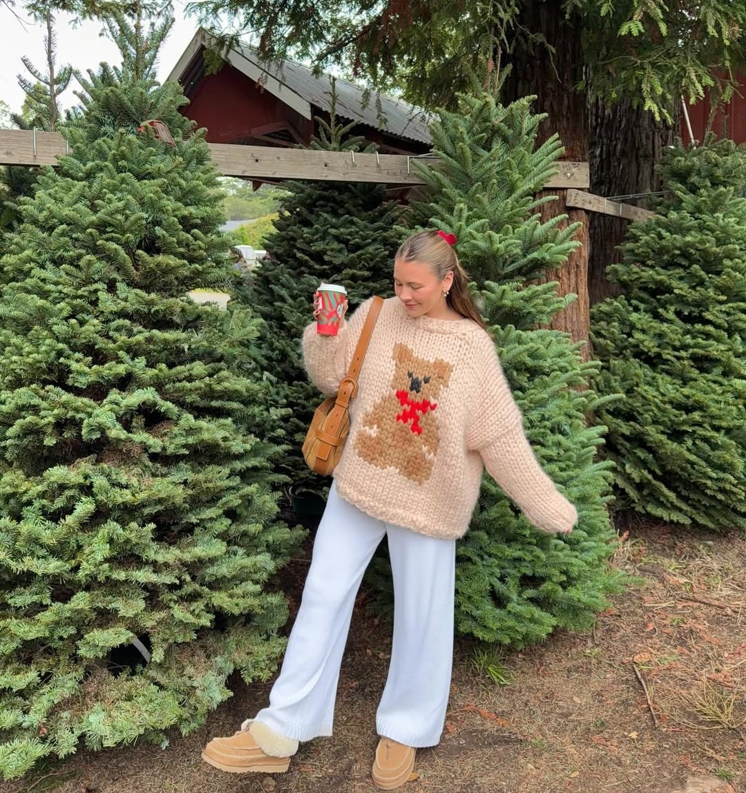 A woman stands among pine trees, wearing a cozy beige sweater with a teddy bear design, white pants, tan slippers, and holding a red cup. She smiles in a festive outdoor setting—perfect to celebrate Swiftmas in style and gather fun Swiftmas ideas.