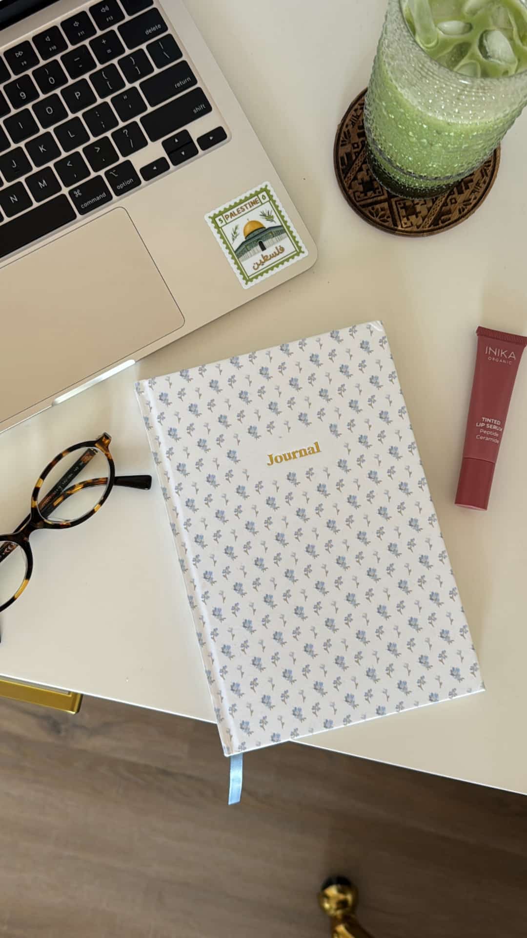 A journal with a blue floral cover lies on a desk beside a laptop, glasses, a small sticker, lip balm, and a green drink—perfect for starting your New Year's journaling habit like your favorite lifestyle YouTubers.
