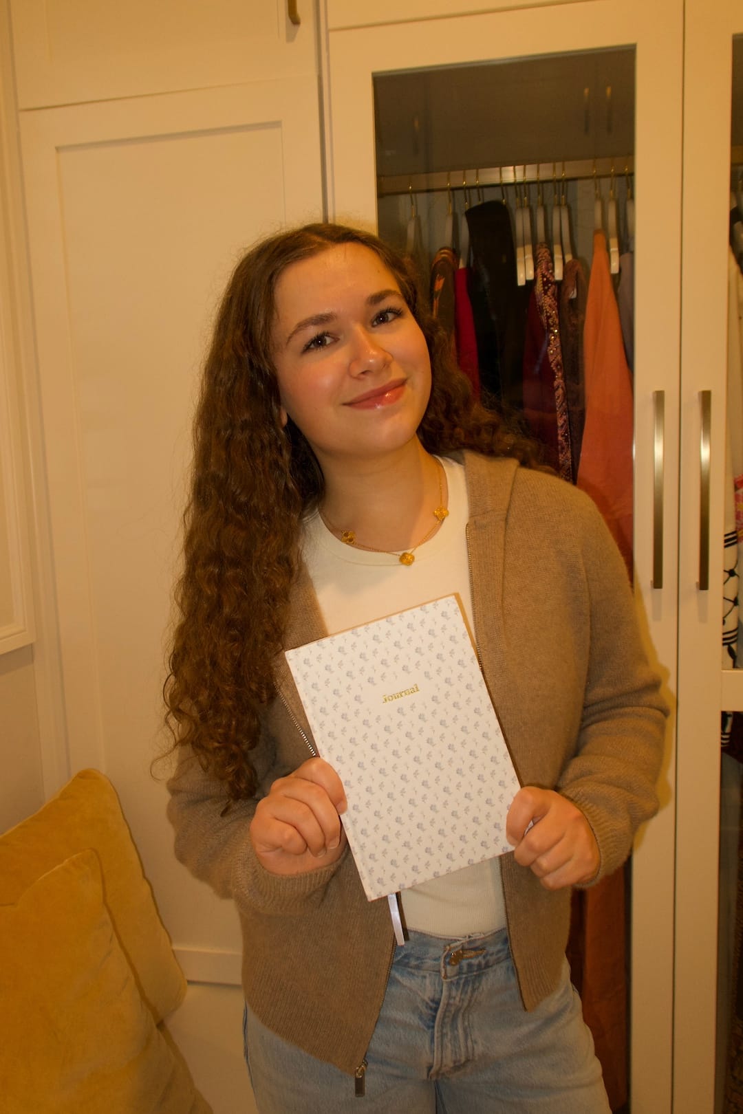 A young woman with long curly hair stands in front of an open wardrobe, smiling and holding a patterned journal—ready to embrace her New Year's journaling habit. She wears a beige cardigan, white shirt, and jeans.