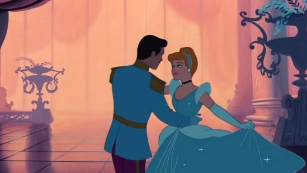 A prince and Cinderella dance together in a grand, softly-lit ballroom—a scene straight from beloved childhood flicks. Cinderella wears a light blue gown and gloves, while the prince is in a blue uniform amid elegant decor and large vases.