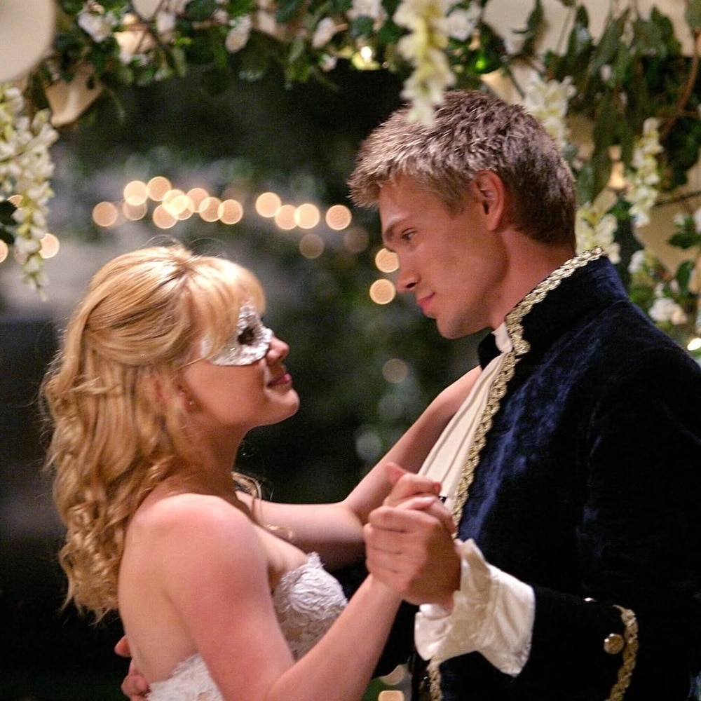A young couple dressed in formal attire dances under a canopy of flowers and lights. Like a scene from romantic comedies, the woman in a white mask and strapless dress and the man in a velvet suit with gold trim gaze into each other's eyes.