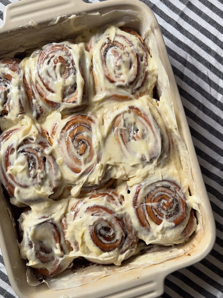A close-up of a baking dish filled with freshly baked cinnamon rolls generously covered in creamy white icing, placed on a black and white striped cloth.