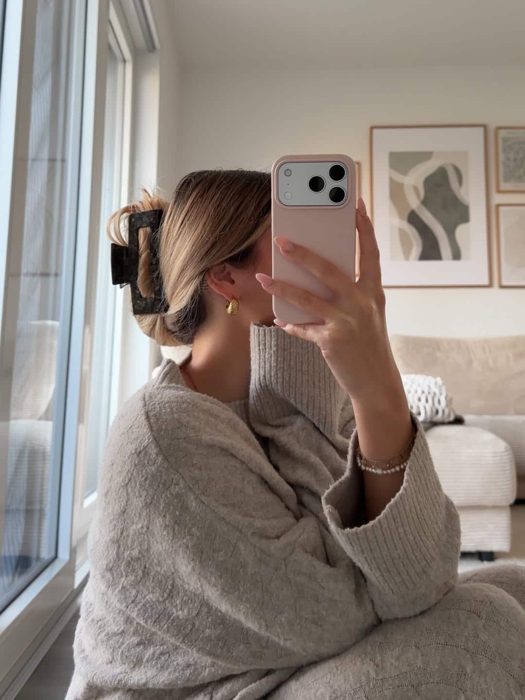 A woman in a cozy beige sweater sits by a window, taking a mirror selfie with her phone. Her hair is tied up with a claw clip, and abstract art hangs on the wall behind her in this modern living room—perfect for styling tips inspiration.