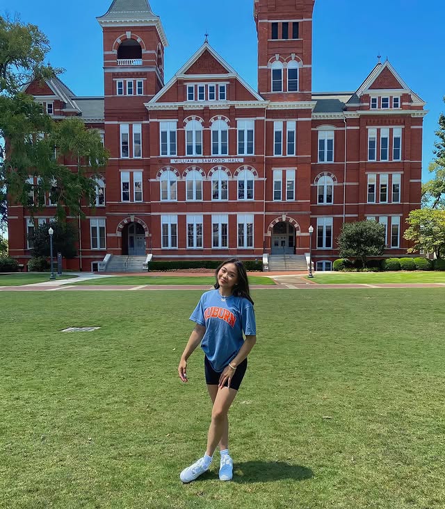 A young woman stands smiling on a large grassy lawn, wearing an Auburn T-shirt and shorts. Behind her is the historic red-brick Samford Hall building at Auburn University, capturing the excitement of college application season under a clear blue sky.