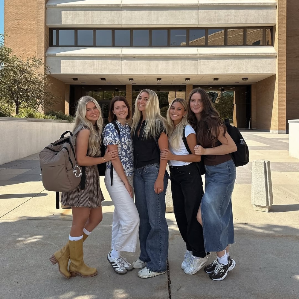Five young women stand together smiling outside a modern building, each wearing casual outfits and backpacks, suggesting theyre students. It’s sunny and they appear happy, posing closely for the photo.