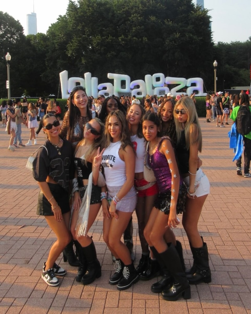 A group of nine young women pose and smile in front of a large Lollapalooza sign at an outdoor festival, with people and trees in the background on a sunny day.