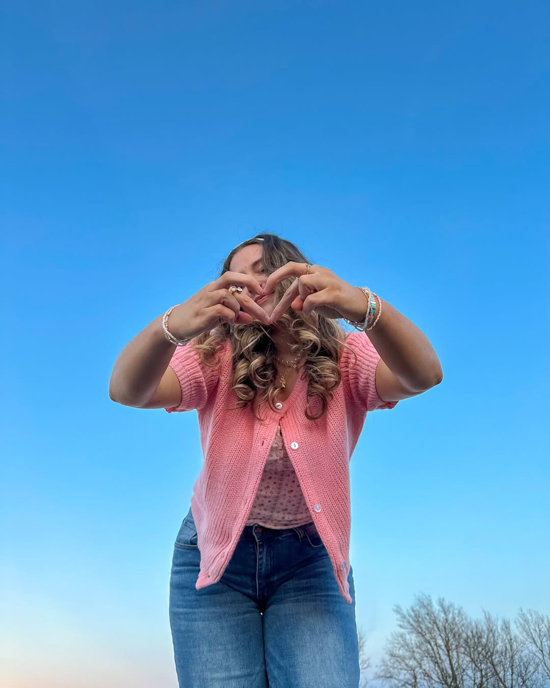 A woman wearing a pink top and blue jeans stands outdoors against a clear blue sky, forming a heart shape with her hands in front of her chest—a reminder to stop comparing love life and embrace your own unique journey. Trees are visible in the lower right corner.