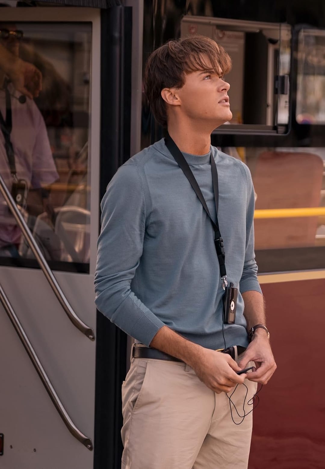 A young man wearing a light blue long-sleeve shirt and beige pants stands outside a bus, holding earphones and a device on a lanyard around his neck, looking thoughtfully upwards, reminiscent of Conrad Fisher on Valentine's Day.