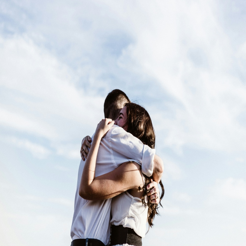 Two people are embracing tightly outdoors under a bright, partly cloudy sky. The scene suggests warmth and affection, offering comfort that can be essential after a break up or when supporting a hurting friend.
