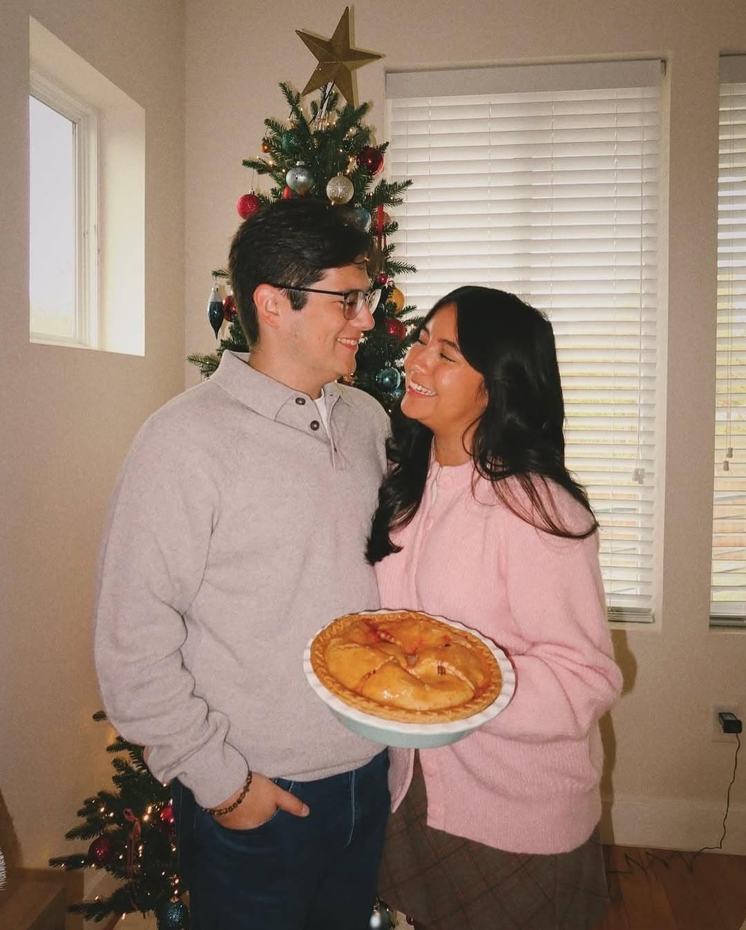 A smiling couple stands in front of a decorated Christmas tree. The man, savoring his holiday crush, holds a pie on a plate as they gaze at each other warmly in cozy sweaters, sunlight streaming through the blinds behind them.