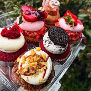 A close-up of assorted cupcakes in a clear plastic tray, topped with frosting, strawberries, Oreo cookies, nuts, and red velvet crumbs, with greenery blurred in the background.