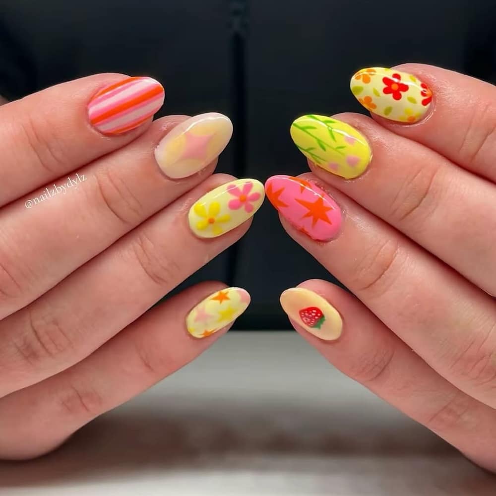 Two hands with almond-shaped nails feature colorful nail art, including flowers, stripes, a strawberry, and leaf designs in vibrant shades—perfect inspiration for the best spa day or adding fun to your at-home spa day.